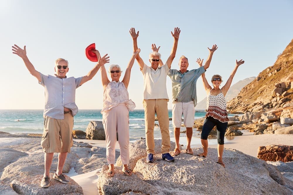 Un grupo de ancianos disfrutan en una playa alzando los brazos y manos hacia arriba. 