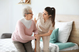 Una cuidadora ayuda a levantarse de la cama a una anciana que porta un bastón.