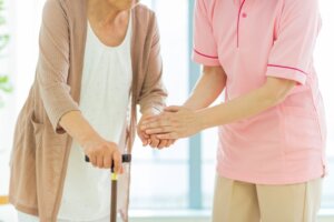 Una cuidadora ayuda a caminar a una anciana que sostiene un bastón en casa.