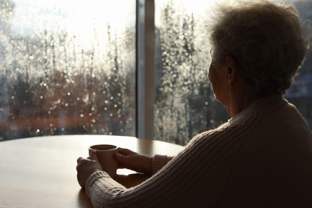 Una anciana mira por la ventana de su casa cómo llueve en el exterior. 