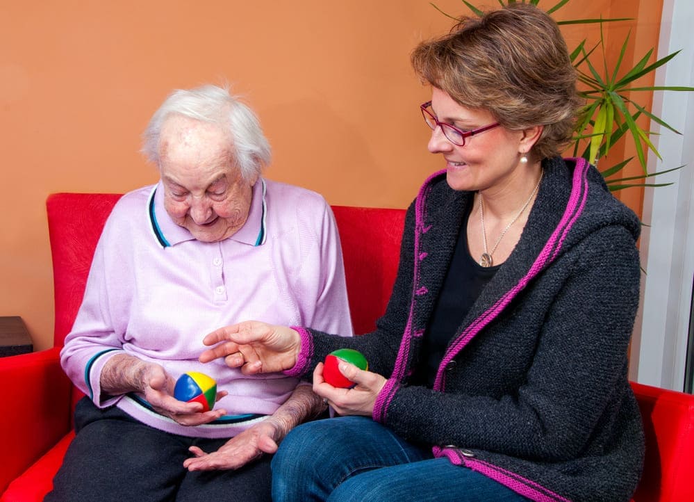 Una cuidadora sostiene una pelota de colores de juguete junto con una señora mayor que sostiene otra mientras ríen. 