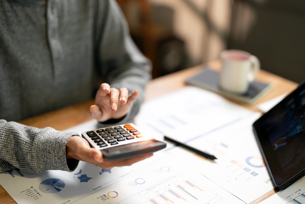 Un plano de una mujer cogiendo una calculadora junto a unos papeles en una mesa y una taza de café. 