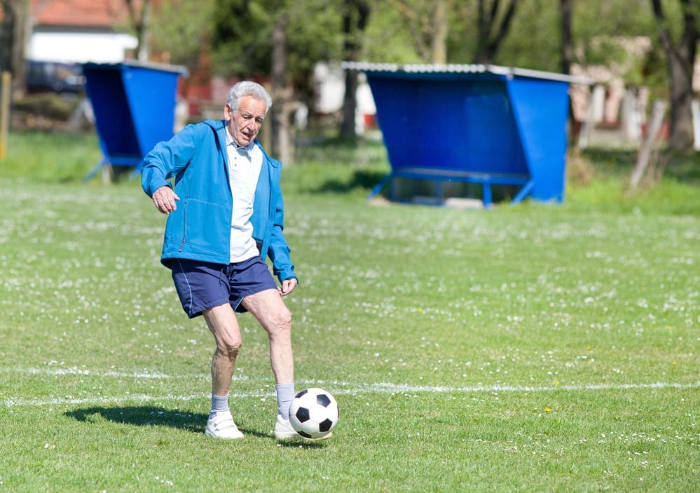 Un señor mayor jugando al fútbol andando en un campo con césped pasa la pelota a un compañero. 