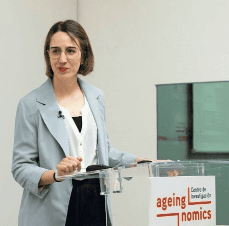 Una imagen de una psicóloga que viste con pelo corto y gafas en un atril en un acto público. 