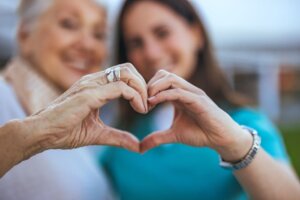 Una chica cuidadora junto a una señora mayor crean con sus manos un corazón.