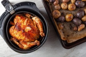 Un pollo entero cocinado en una freidora de aire, junto a una bandeja de patatas.