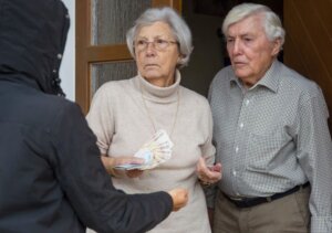 Una anciana y un anciano charlan con una persona que es un estafador. Ellos llevan billetes en la mano.