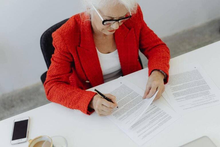 Una señora mayor con una chaqueta americana roja firma un papel.