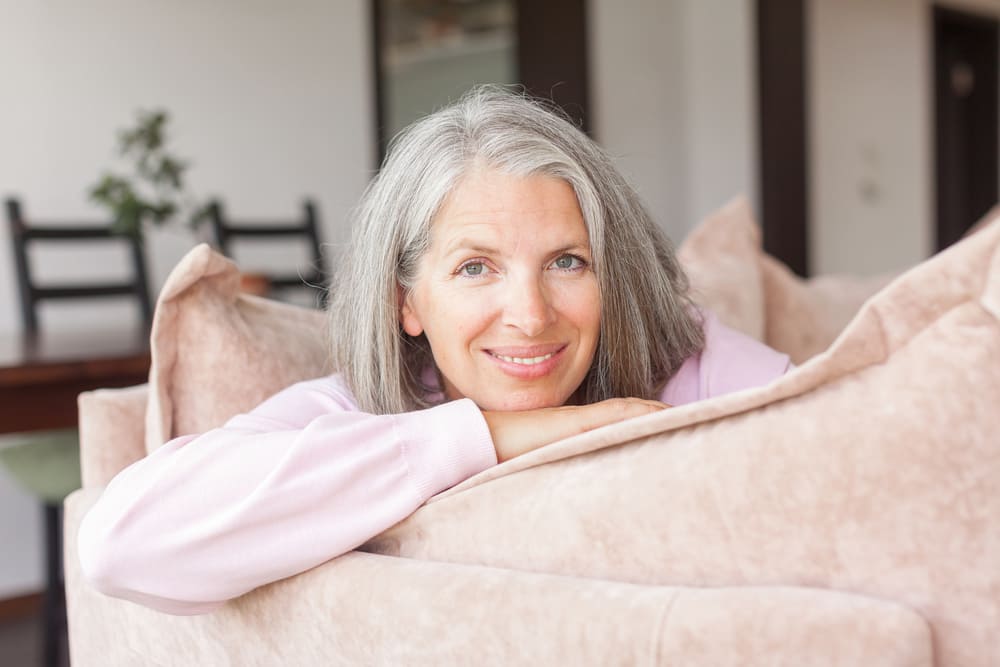 Una señora de unos 60 años mira a cámara mientras está sentada en su sofá sonriendo. 