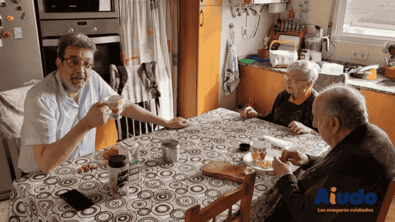 Un señor mayor y una señora mayor desayunando junto a un cuidador hombre en la mesa de una cocina. 