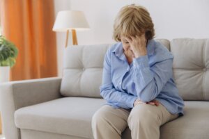 Una cuidadora sentada en el sofá de una casa con una mano en la cabeza como señal de cansancio.