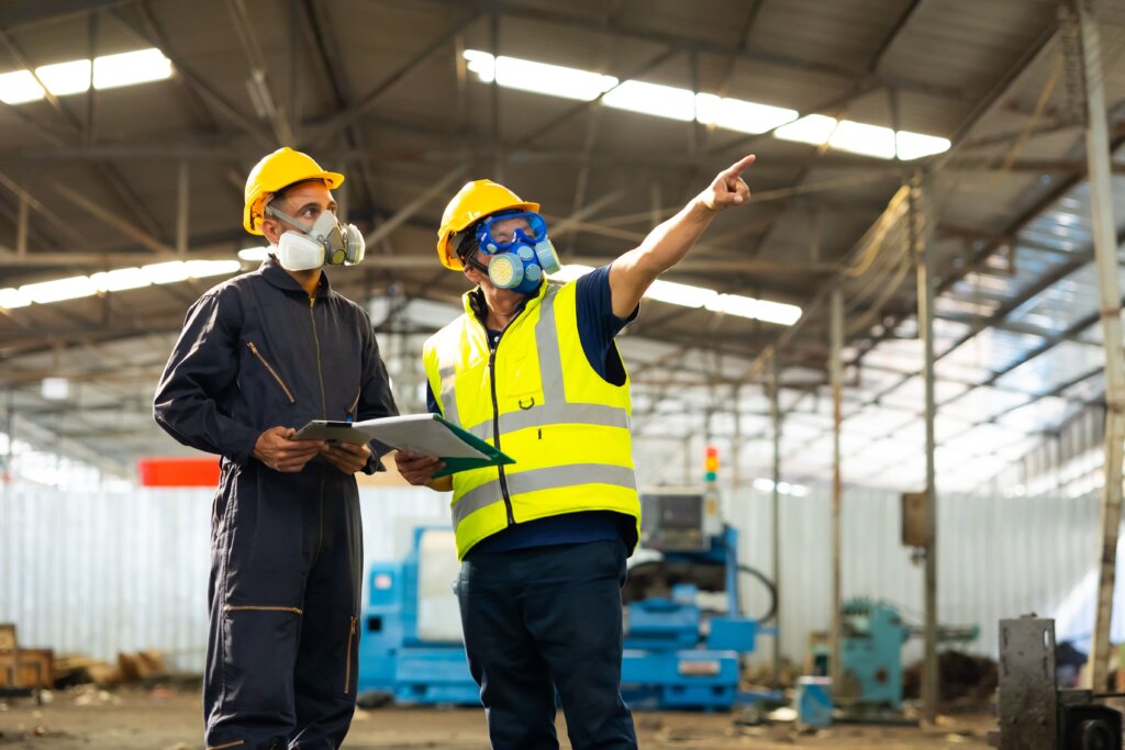 Jubilación de actividades peligrosas Dos operarios en una nave con protecciones: chaleco de seguridad, ropa de seguridad, cascos y mascarillas profesionales haciendo revisiones