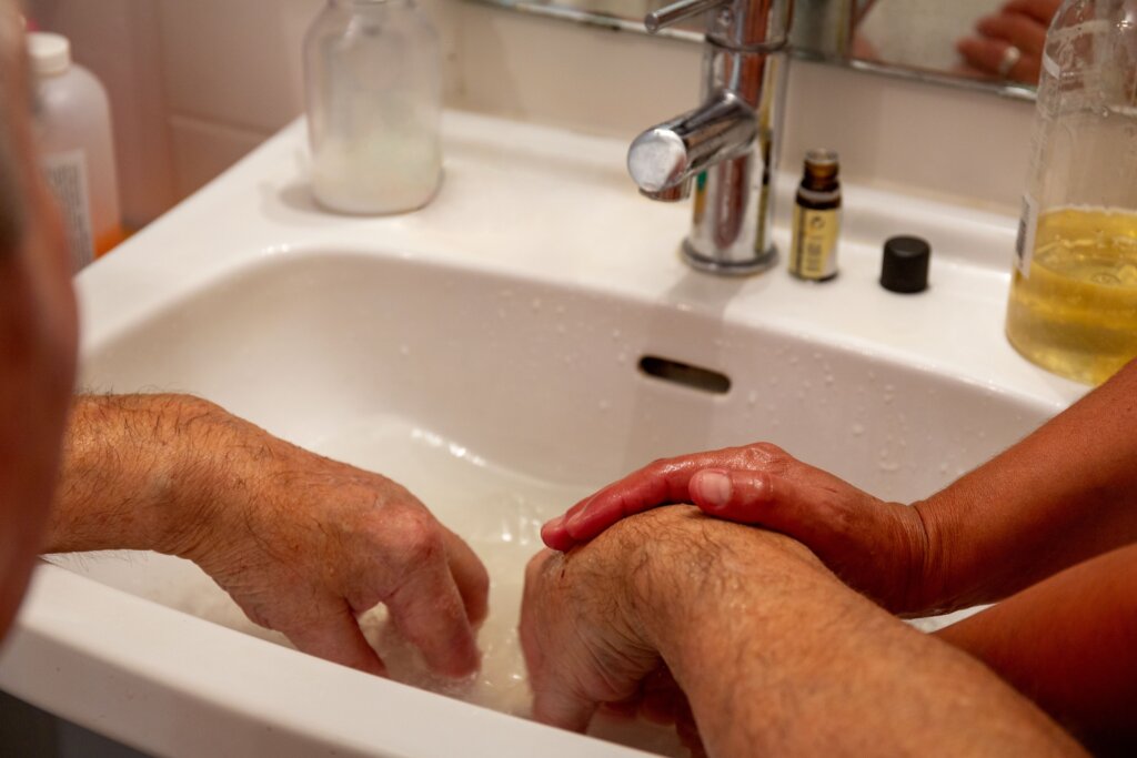 Manos de una persona mayor sobre la pila de un lavabo siendo lavadas con ayuda de una cuidadora con cariño y paciencia 