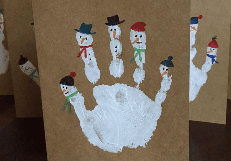 postal de manualidad en la que se estampa la mano con pintura blanca y en los dedos se dibujan muñecos de nieve. 