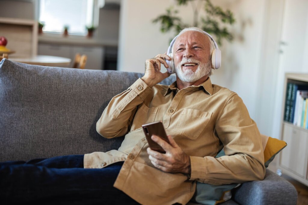 Podcast para persona mayor Persona mayor escuchando con los cascos y el móvil un podcast sonriente desde el sofá