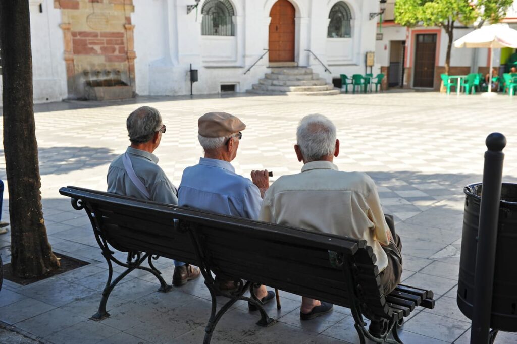 Envejecimiento de la población Tres hombres de la tercera edad sentados en el banco de una plaza de espaldas a la cámara conversando.