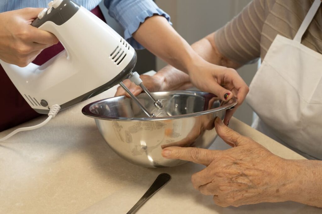 Envejecer en casa Manos de cuidadora con una batidora de varillas, mientras la persona mayor participa sosteniendo el bowl.