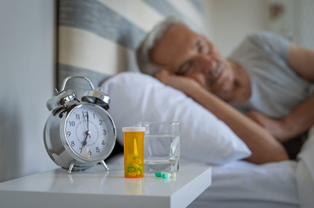 Somnoliencia en personas con Alzheimer Persona mayor en la cama durmiendo, en su mesita tiene un despertador, un vaso de agua y medicación