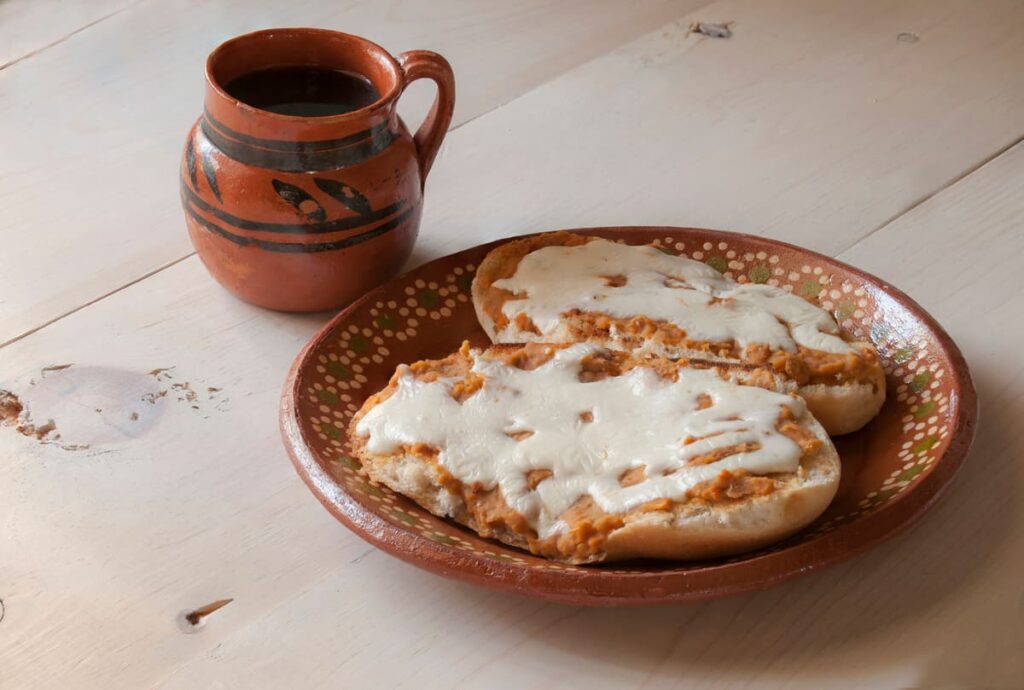 Meriendas de los abuelos plato de barro con un mollete a la mitad con manteca y queso junto a una jarra de barro con bebida sobre la mesa