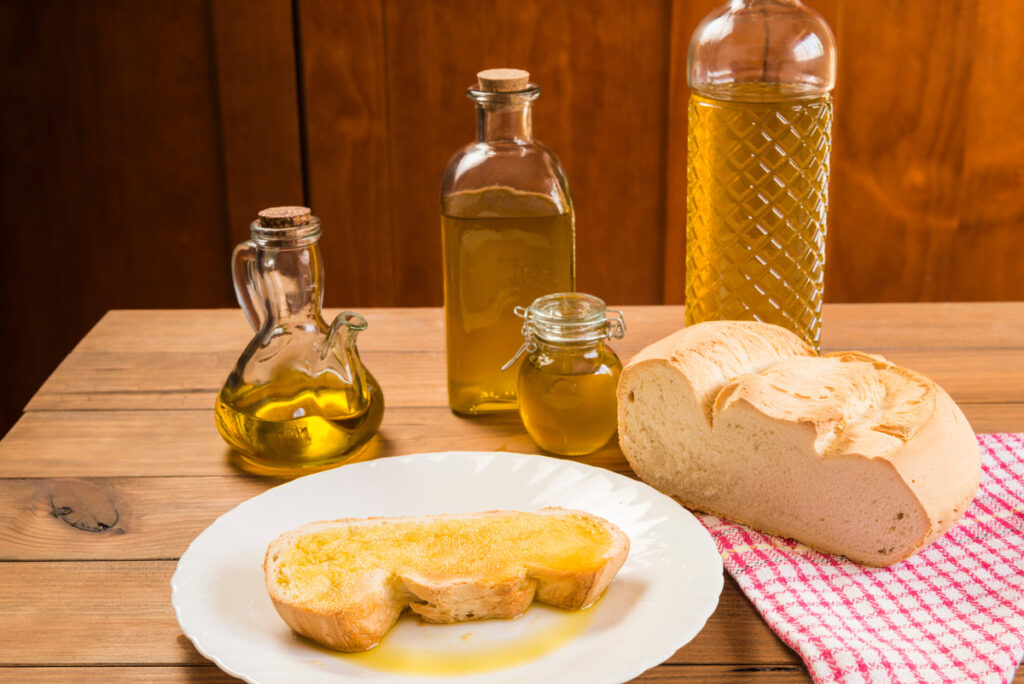 Meriendas de los abuelos Bodegón de una rebanada de pan con aceite de oliva virgen extra sobre un plato blanco, junto a botellas y recipientes llenos de aceite, sobre una mesa rústica de madera. merienda tradicional de la cocina mediterránea.