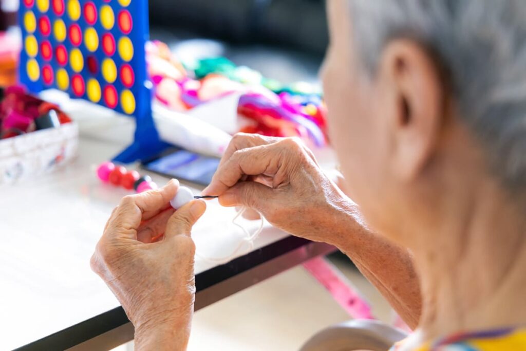 Ejercicios cognitivos persona mayor Anciana realizando un juego cognitivo que consiste en insertar sobre un hilo con una aguja bolas de diferentes colores.