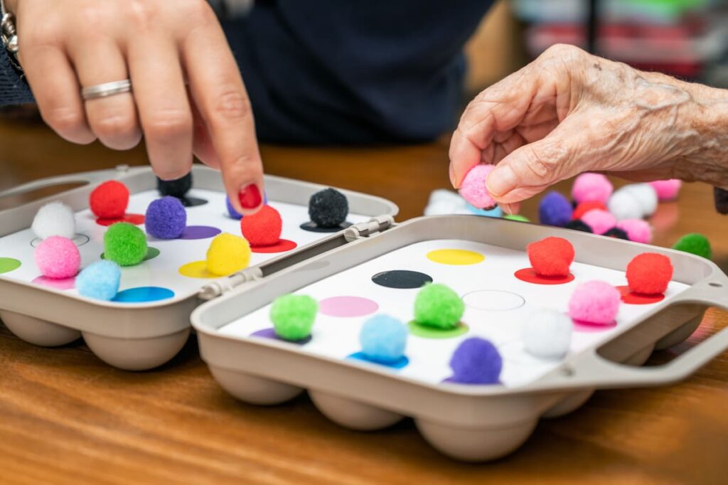 Juegos manuales para personas con Alzheimer Mano de la cuidadora indicando y la persona mayor cogiendo una bola de lana realizando un juego manual sensorial. Se trata de una huevera de base, una cartulina con círculos de colores encima y bolas de lana pequeñas de esos colores que deben colocarse sobre las casillas de su correspondiente.